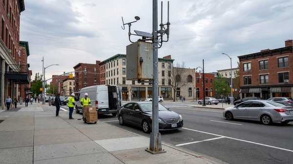 Roadside communications unit mounted on a signal pole overlooking a downtown curb lane with mixed loading and parking activity